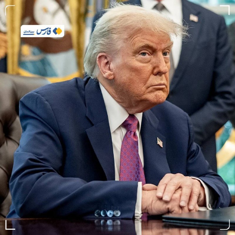 A man with blonde hair in a navy suit and patterned tie sits at a desk with clasped hands, with another suited person standing behind him.