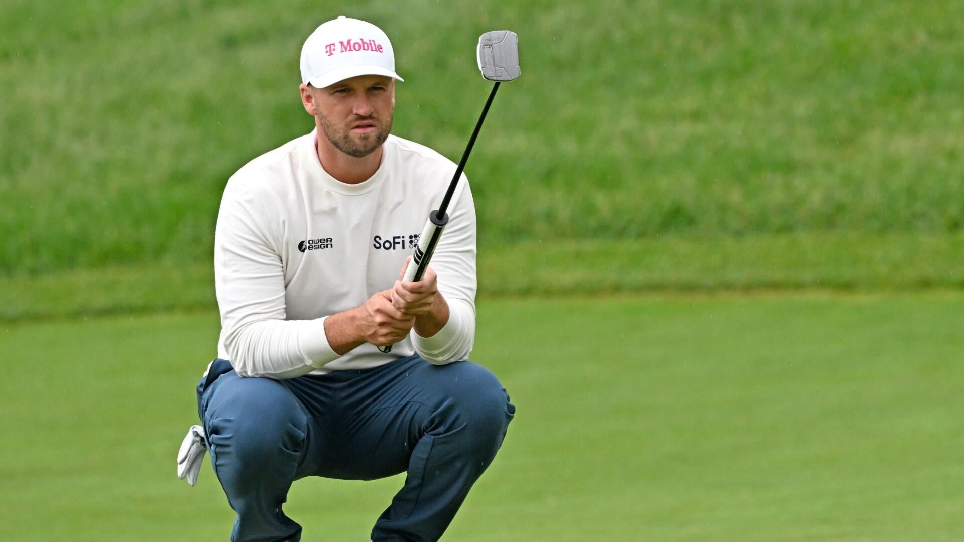 A bearded golfer wearing a white T-Mobile cap and shirt crouches on a green, holding up a putter to study the grass.