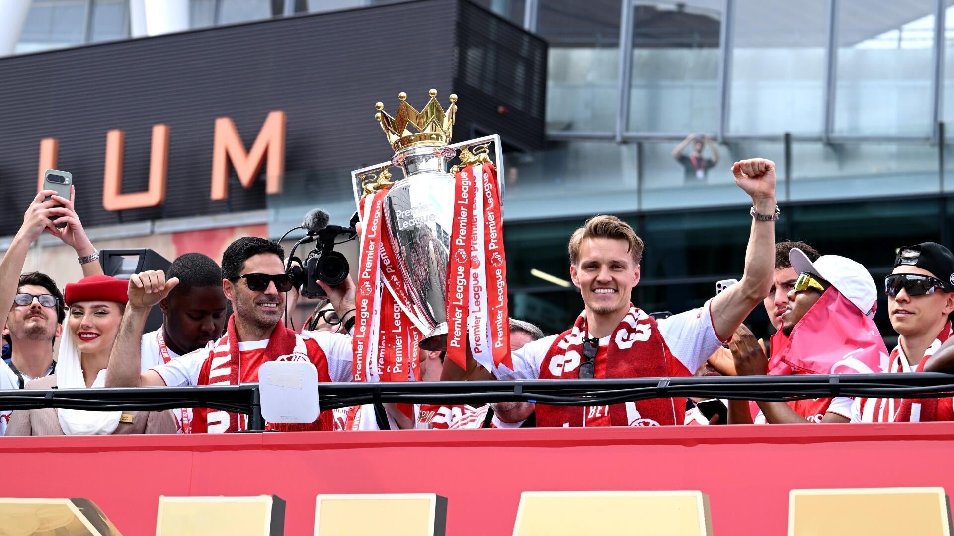Celebrating on a parade bus, smiling people wearing red and white scarves hold up a large silver Premier League trophy topped with a golden crown, surrounded by cameras and a crowd.