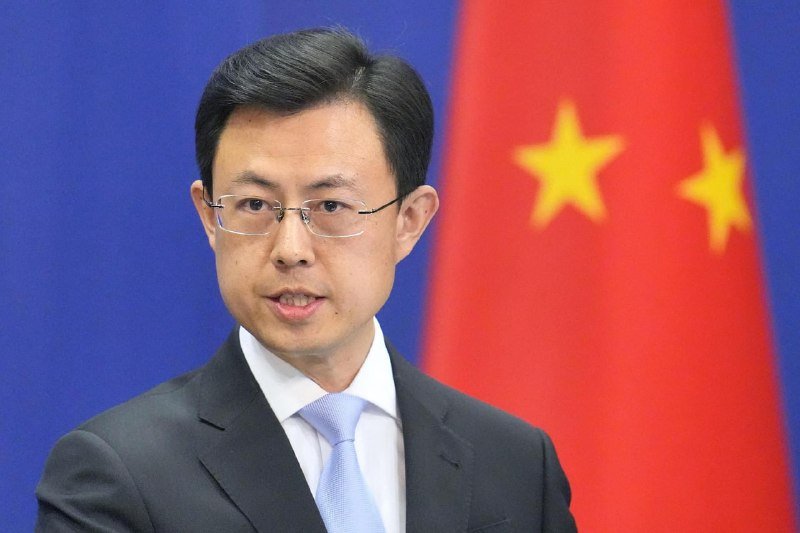 A man in a dark suit and light blue tie speaks at a podium, with a blue backdrop and a red Chinese flag with yellow stars visible behind him.
