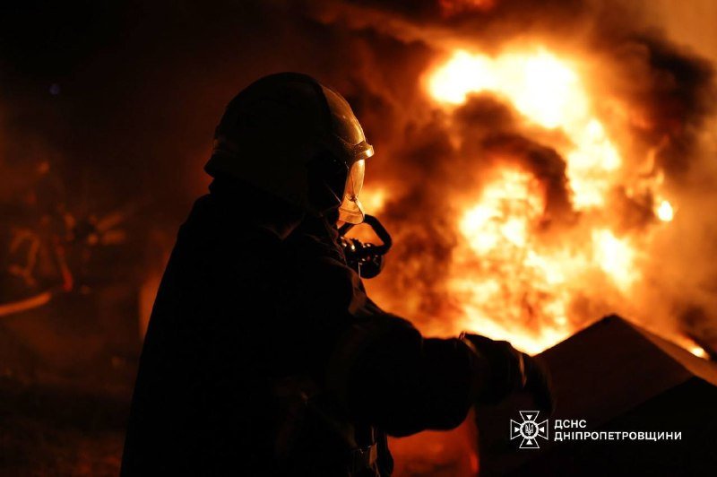 A silhouetted firefighter in a helmet directs a hose at large orange flames at night, with a logo reading "ДСНС Дніпропетровщини" in the corner.