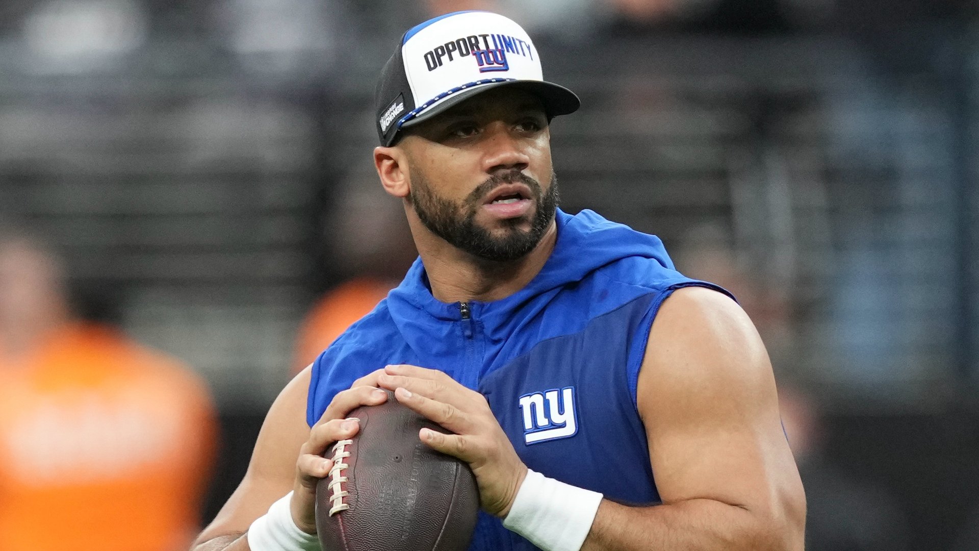A bearded football player wearing a blue New York Giants sleeveless hoodie and a white "Opportunity" cap holds a football in a throwing stance.