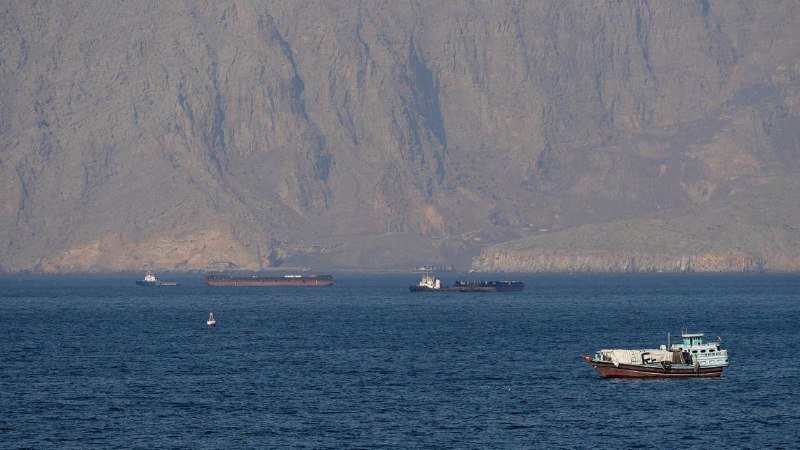 Several cargo ships and a smaller vessel float on calm blue water, with a massive rocky mountain range rising steeply in the background.