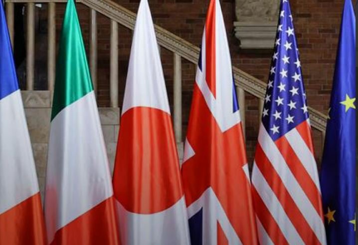 Five flags—the US, UK, Italy, France, and EU—stand displayed in a row against a stone staircase backdrop.