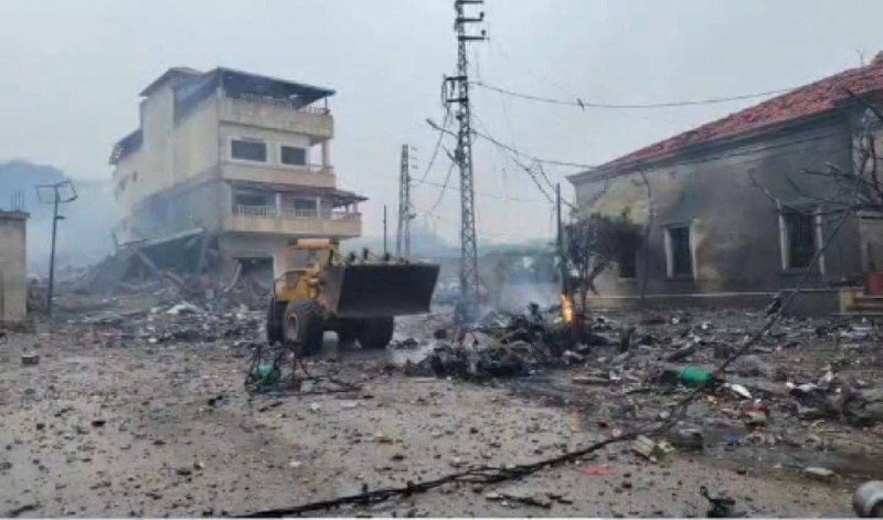 A yellow front-end loader operates amid rubble and scattered debris between damaged buildings, with a small fire burning and smoke rising in the background.