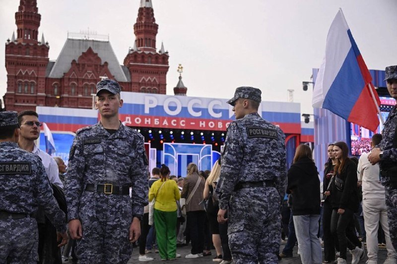 Uniformed personnel with "Росгвардия" patches stand guard near a Russian flag and a stage reading "РОСС ДОНБАСС-НОВ," with a red historic building in the background.
