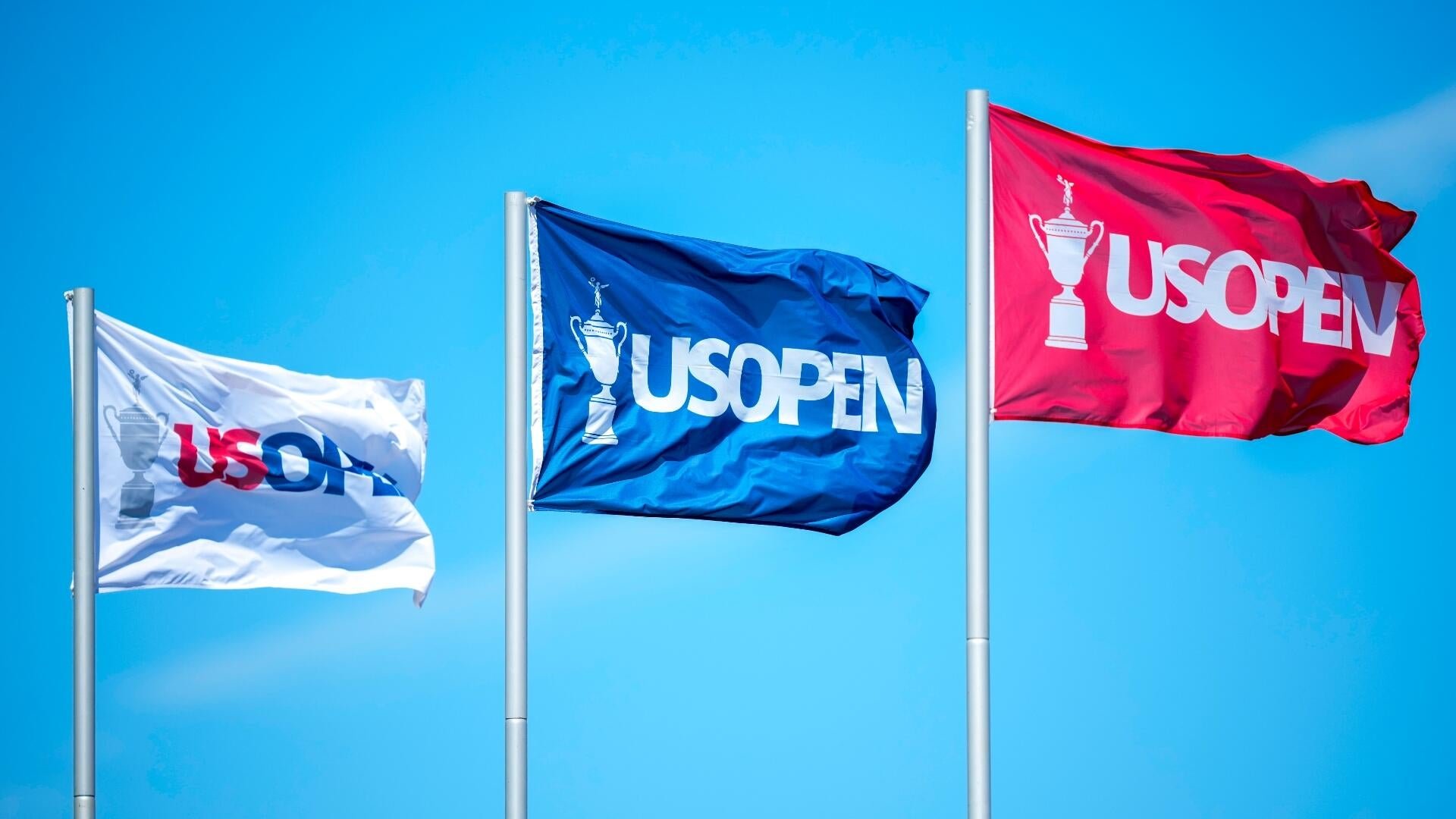 Three US Open flags (white, blue, and red) displaying a trophy emblem flutter on flagpoles against a clear blue sky.
