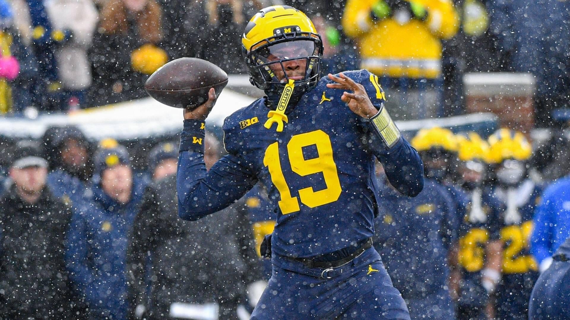 A football player wearing a navy and yellow #19 jersey and helmet throws a pass as snow falls during a game, with spectators and teammates visible in the background.