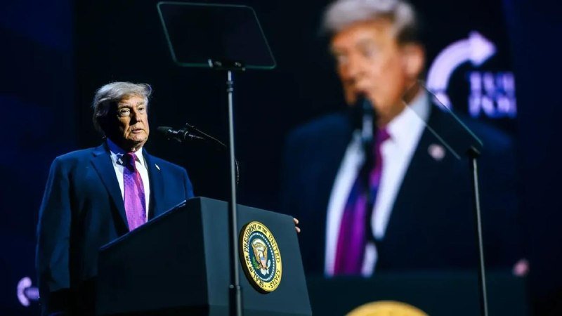 A man with blonde hair in a dark suit and purple tie speaks at a podium bearing a presidential seal, with a large blurred projection of himself visible behind him on stage.
