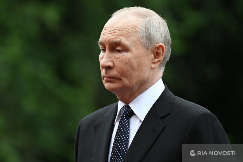 A gray-haired man in a dark suit and polka-dot tie stands outdoors with a serious expression against a blurred green background, with a RIA Novosti watermark visible.
