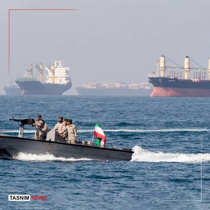 Four uniformed personnel operate a small speedboat flying the Iranian flag across open water, with large cargo ships visible in the hazy background.