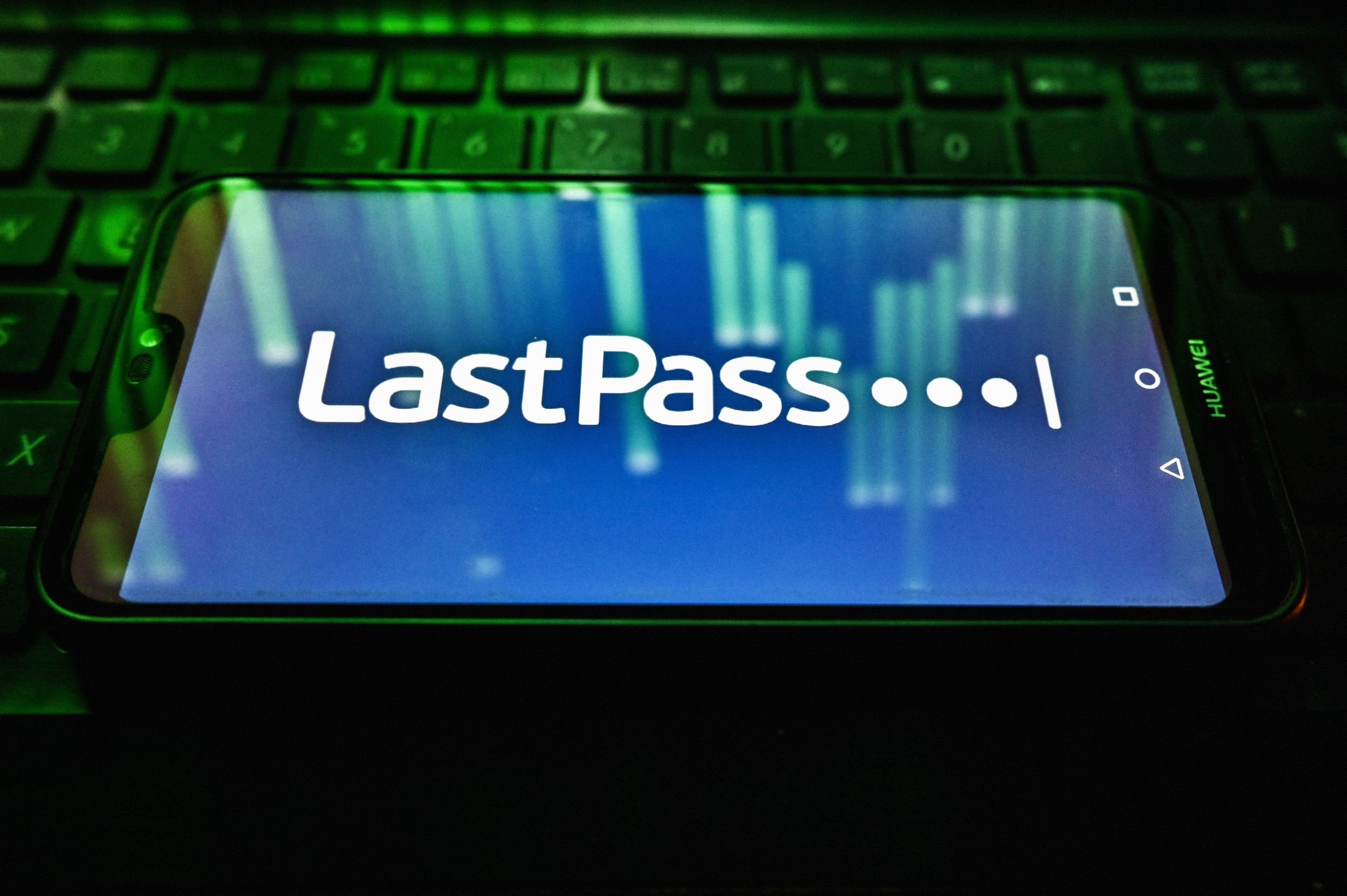 A Huawei smartphone displays the LastPass logo on a blue background, resting on a keyboard illuminated in green light.