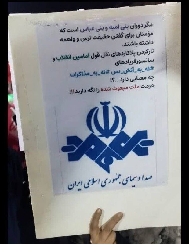 A printed poster with Persian text and a stylized emblem featuring crossed swords and a crown-like symbol is held up against a background, with a hand making a peace sign visible at the bottom.