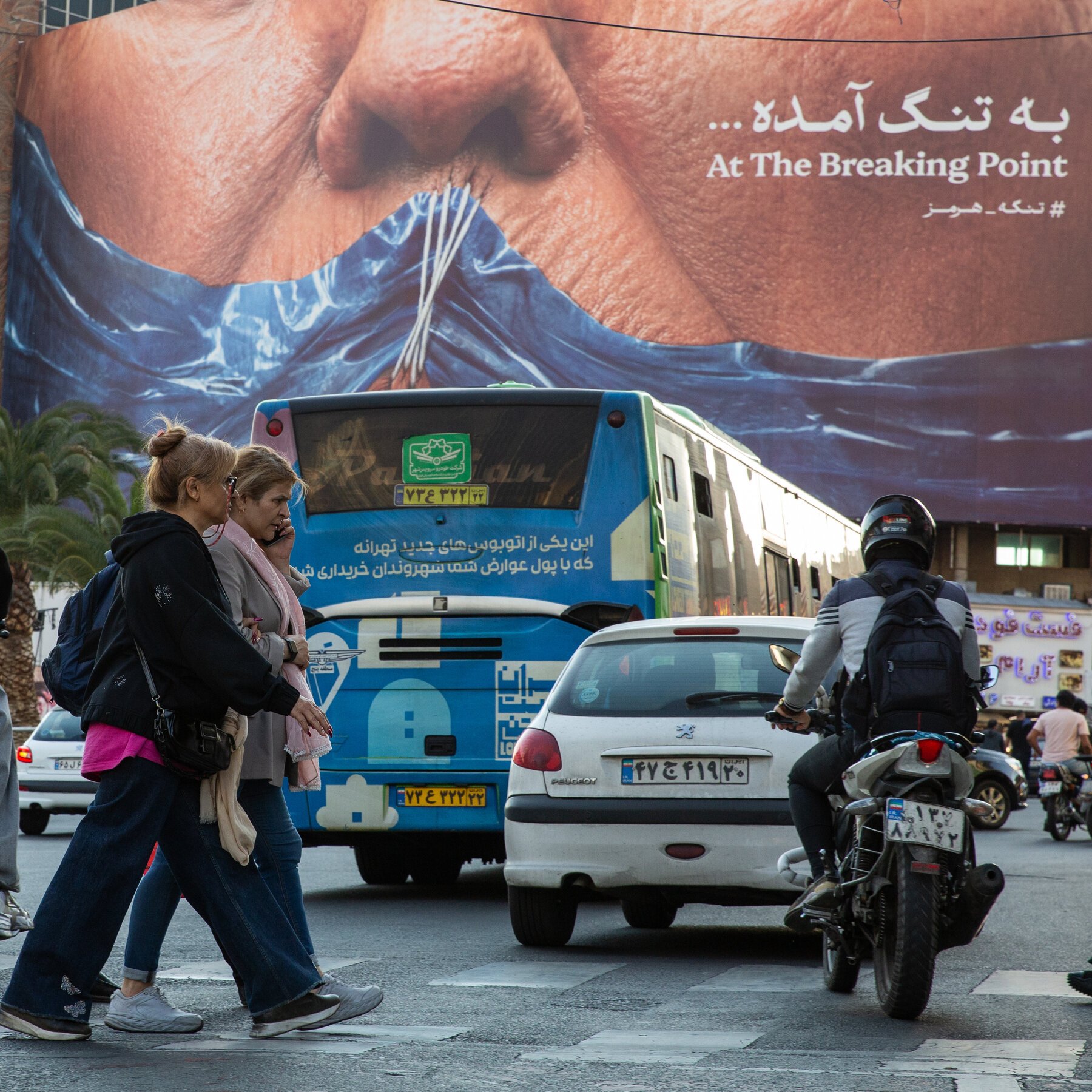 Two women cross a busy street in front of a blue bus, white car, and helmeted motorcyclist, beneath a large billboard reading "At The Breaking Point" in English and Persian.