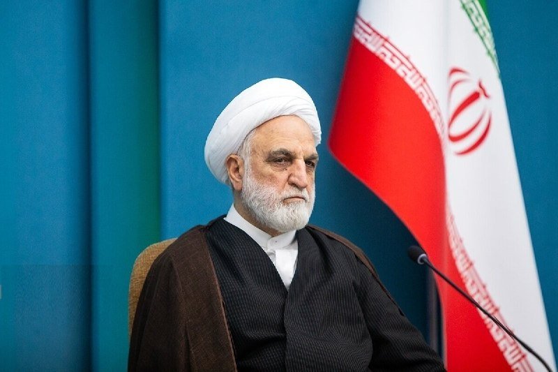 A bearded cleric wearing a white turban and black robe sits before a microphone, with the Iranian flag displayed behind him.