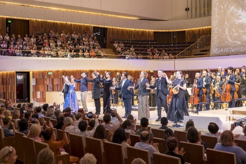 Singers and performers in formal attire take a bow on a curved stage before a full, applauding audience inside a modern concert hall.