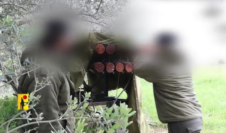 Two blurred figures in military attire stand beside a covered weapon system with cylindrical launch tubes, set in a wooded outdoor area.