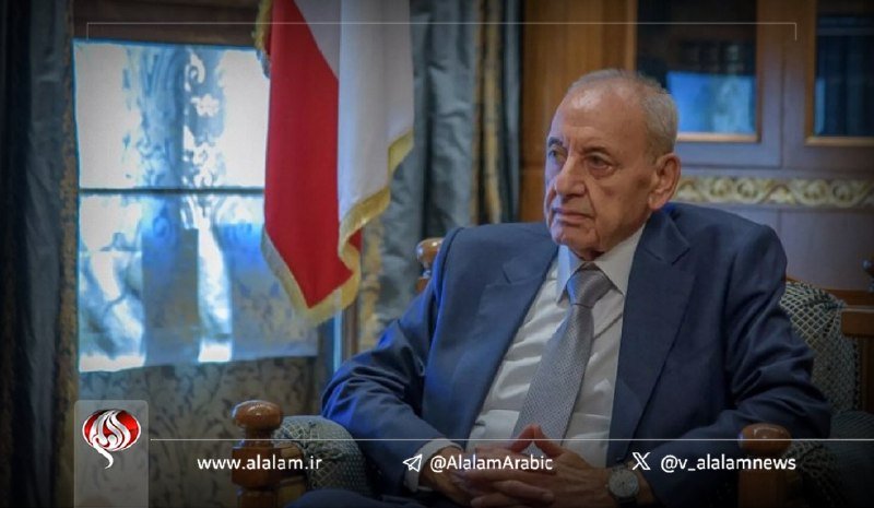 An older man in a blue suit and gray tie sits in a chair beside a red, white, and black flag during what appears to be an interview.