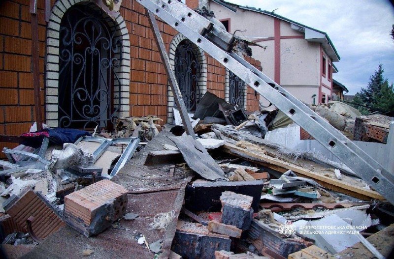 Debris, broken bricks, twisted metal beams, and damaged building facades are scattered across the ground in front of a partially destroyed brick structure.
