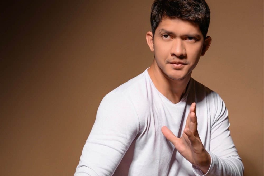 A man in a light gray long-sleeve shirt poses against a brown backdrop, gesturing with one open hand while looking off to the side.