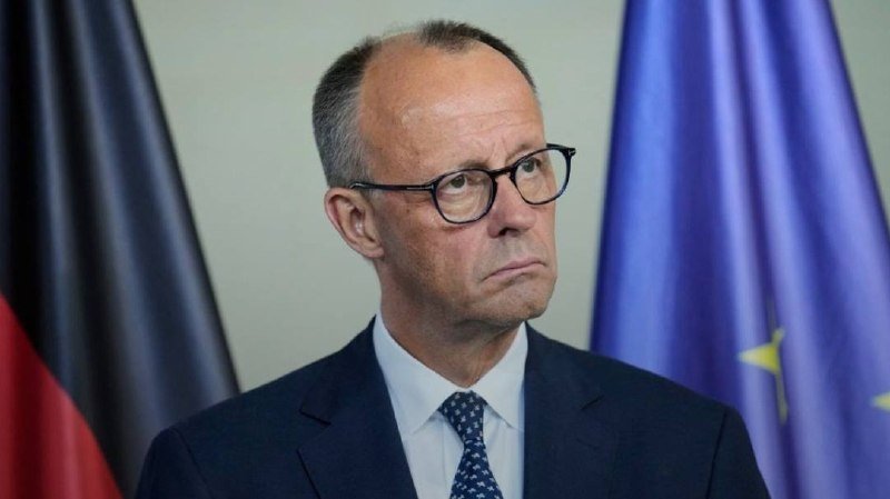 A bespectacled man in a dark suit and patterned tie stands between a German flag and a European Union flag.