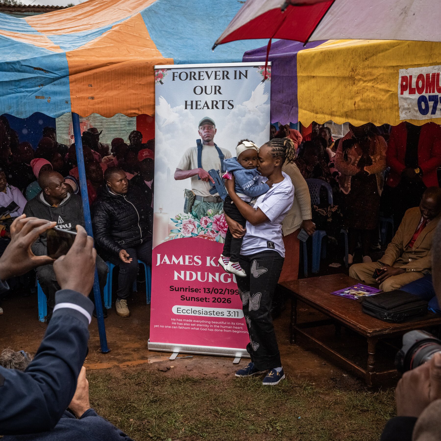 Mourners gather under colorful tents at an outdoor memorial service, where a woman holds a child beside a large portrait banner reading "Forever in Our Hearts."