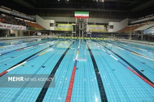 An empty indoor Olympic-sized swimming pool with multiple lanes, Iranian flags, and Arabic script banners displayed in the stands.