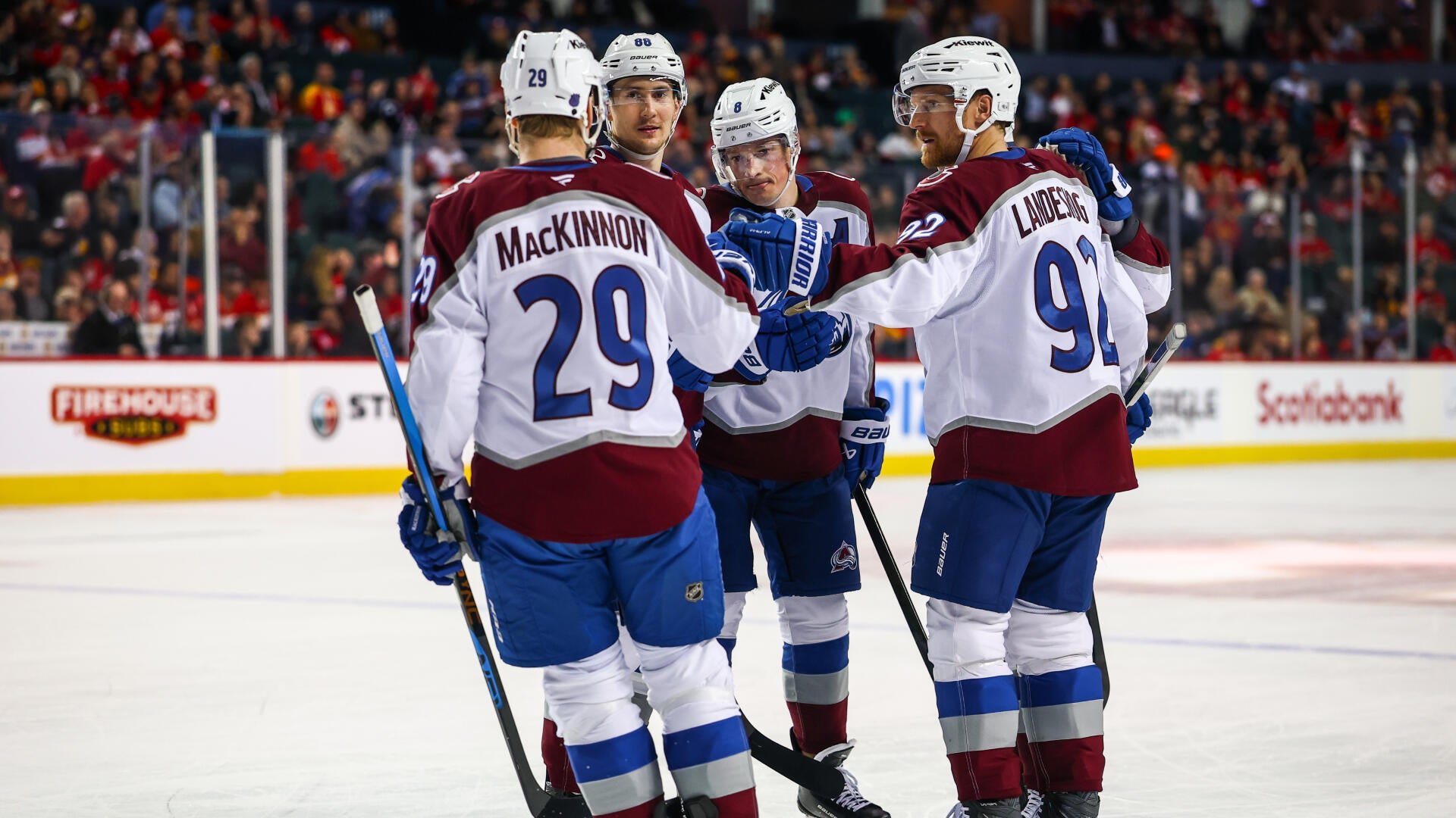 Four hockey players in burgundy, white, and blue uniforms gather on the ice, with one player raising a gloved hand toward another, in front of a crowded arena.