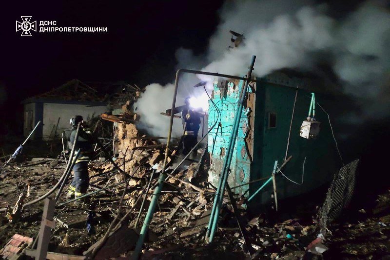 Firefighters work amid the smoldering wreckage of a destroyed building at night, with smoke rising and debris scattered throughout the scene.