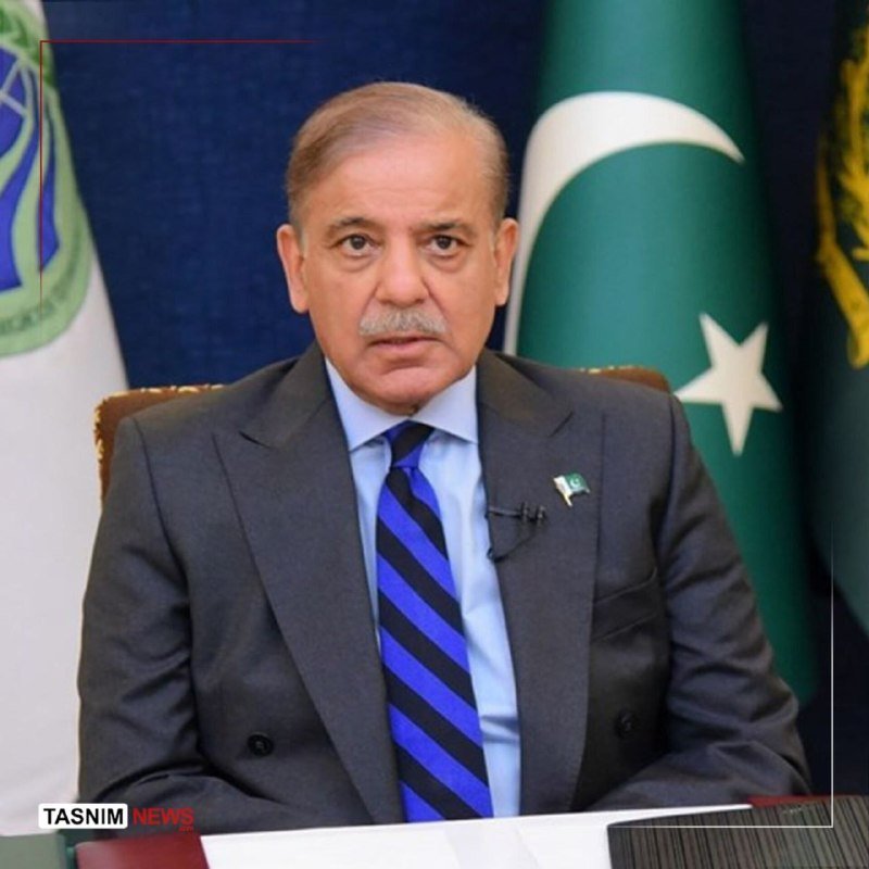 A man in a gray suit and blue striped tie with a lapel microphone sits before flags, with a "Tasnim News" watermark visible.