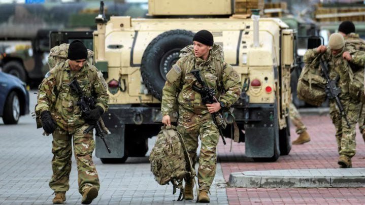Armed soldiers in camouflage uniforms carry rifles and gear while walking on a paved area near a large tan military vehicle.