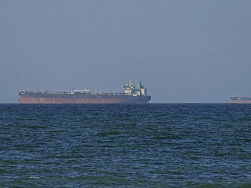 A large red-hulled tanker ship sails across choppy gray-blue waters under a hazy sky, with a second vessel faintly visible on the distant horizon.