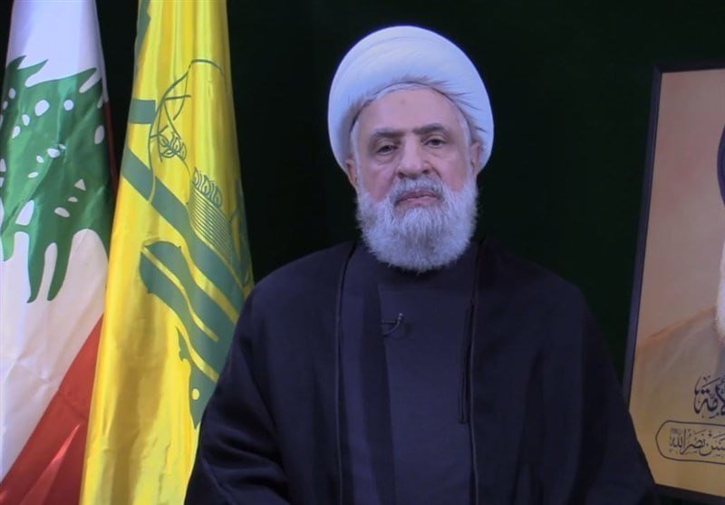 A bearded man in black clerical robes and a white turban speaks, flanked by the Lebanese flag and a yellow Hezbollah flag, with a framed portrait visible on the right.