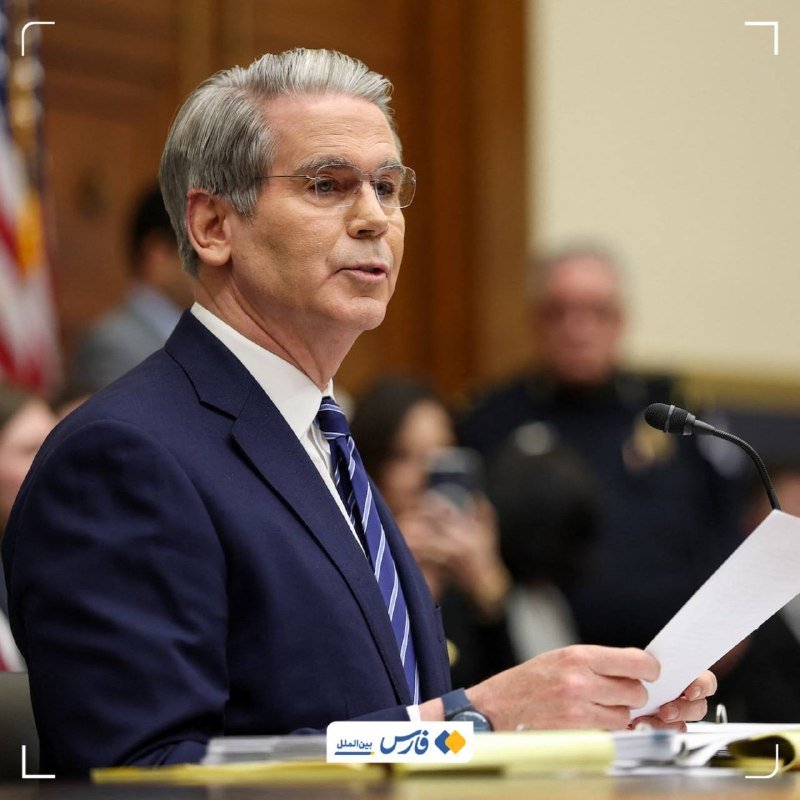 A gray-haired man in a dark suit, white shirt, and blue striped tie speaks into a microphone while holding papers in a formal indoor setting.