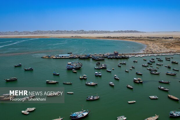 Numerous small boats are anchored in a green coastal waterway near a sandy shoreline under a clear blue sky.