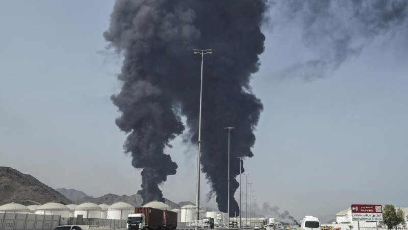 Thick black smoke billows into the sky from an industrial facility containing white storage tanks, with trucks and streetlights visible in the foreground.