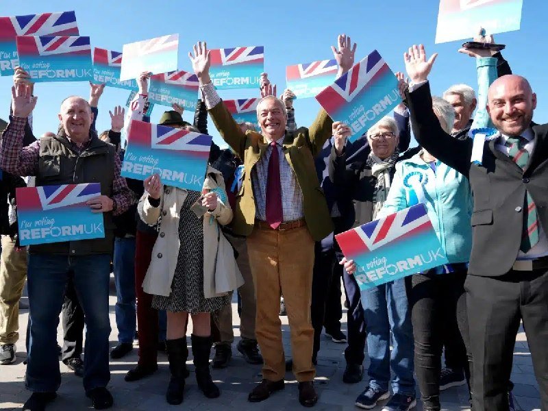 A group of smiling people stand outdoors waving blue "I'm voting REFORM UK" signs featuring the Union Jack flag.