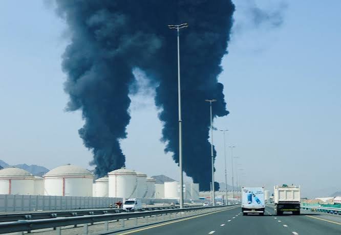 Thick black smoke rises from white cylindrical storage tanks beside a highway as trucks and cars travel down the multi-lane road.