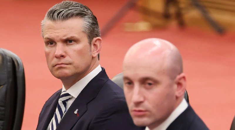 Two men in dark suits sit side-by-side in an ornate, red-carpeted room, looking forward with serious expressions.