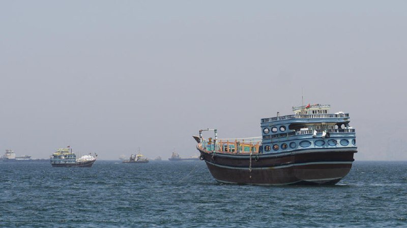 A large multi-deck cargo vessel with a dark hull and blue-trimmed superstructure floats on choppy gray waters, with several smaller ships visible in the hazy distance.