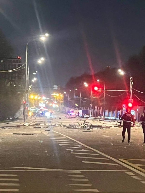 Nighttime street scene showing debris scattered across a road, emergency vehicles, and personnel in reflective gear near traffic signals.