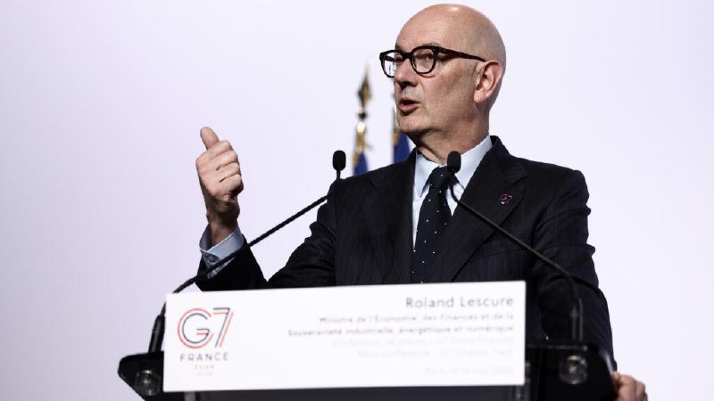 A bald man with glasses speaks at a G7 France podium, gesturing with one hand.