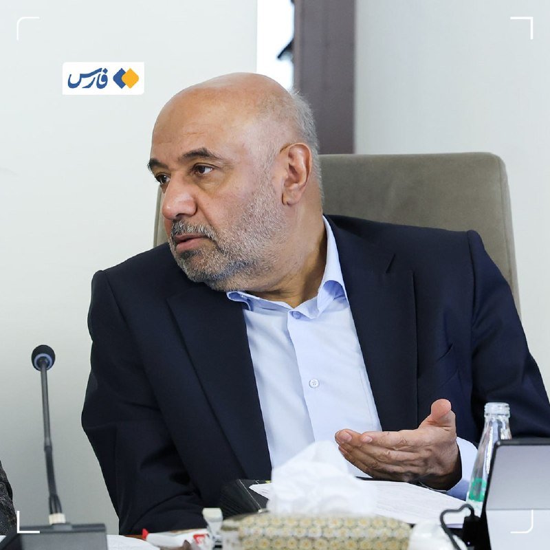 A bald, bearded man in a dark suit and light blue shirt sits at a conference table, gesturing with his hand while speaking.