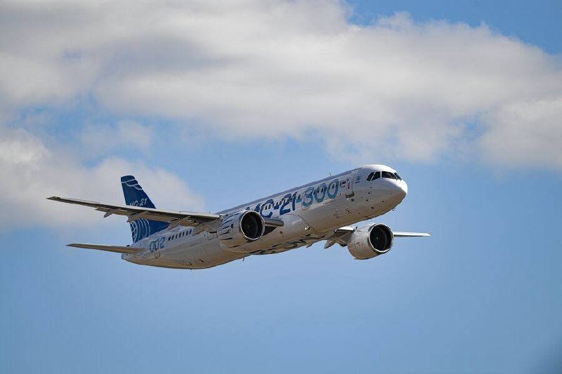 A commercial passenger jet marked "AC21-300" and "002" on its fuselage ascends through a partly cloudy blue sky.