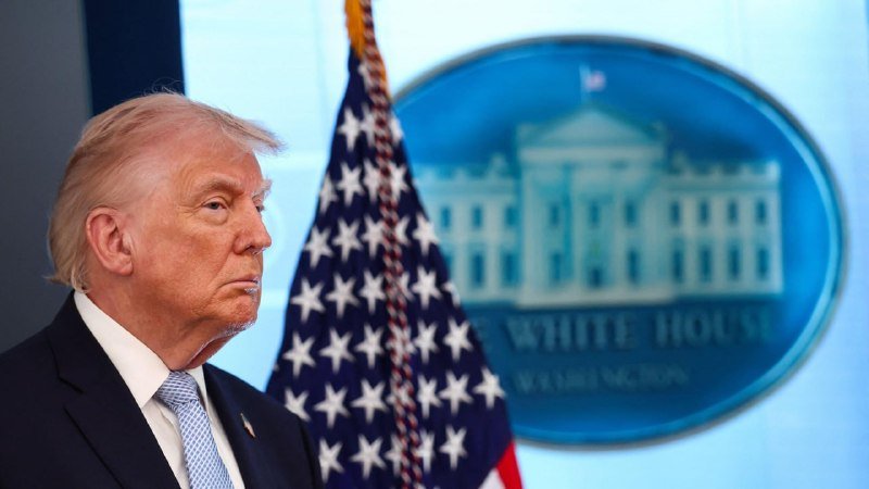 A blonde-haired man in a dark suit and light blue tie stands beside an American flag in front of the White House press briefing backdrop.