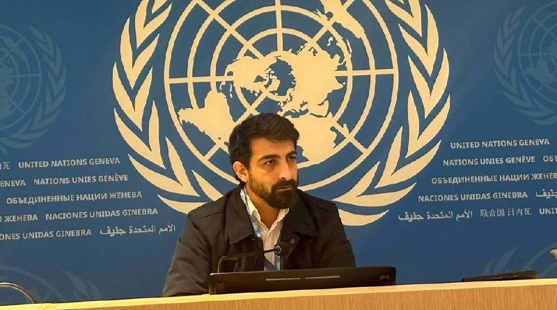 A bearded man in a dark jacket sits at a desk in front of a blue backdrop displaying the United Nations emblem and multilingual "UNITED NATIONS GENEVA" text.