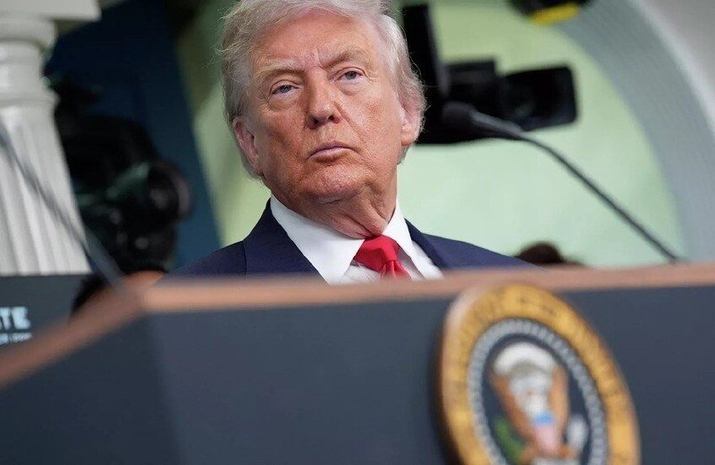 A man in a dark suit and red tie stands at a podium bearing a presidential seal, with a camera and microphone visible in the background.
