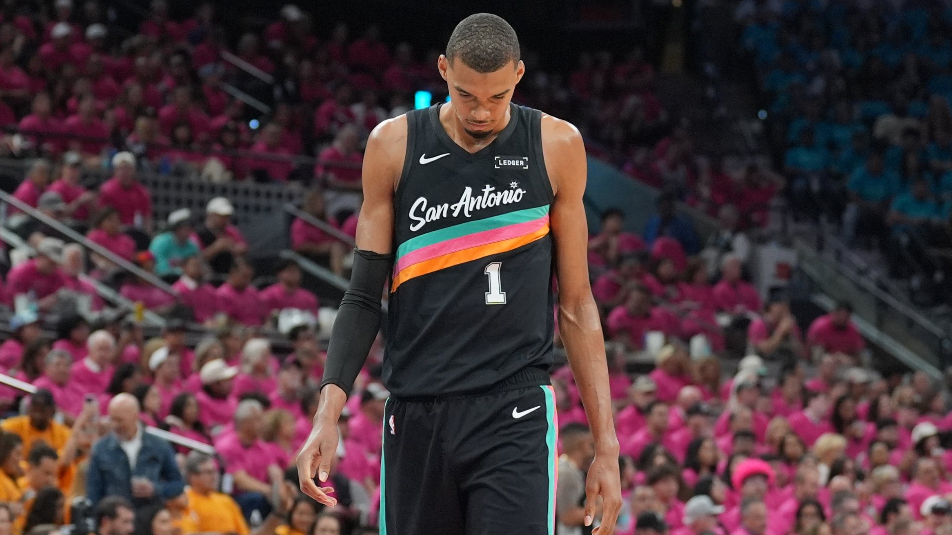 A San Antonio basketball player wearing jersey number 1 stands with his head down on the court, surrounded by a crowd of fans wearing pink.
