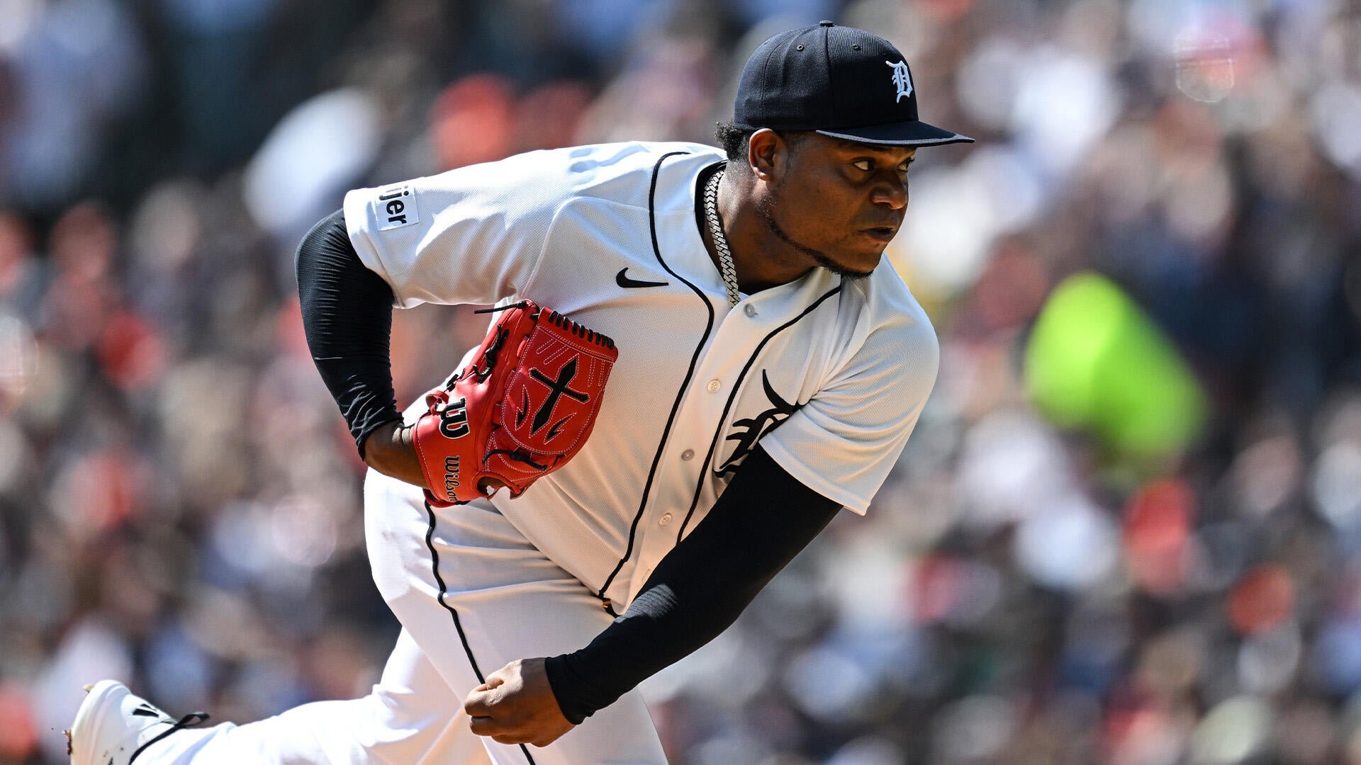 A baseball pitcher in a white Detroit Tigers uniform and navy cap winds up to throw, wearing a red glove, with a blurred crowd in the background.