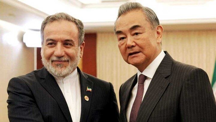 Two older men in dark suits stand close together smiling for a photo indoors, one wearing a small flag pin on his lapel.